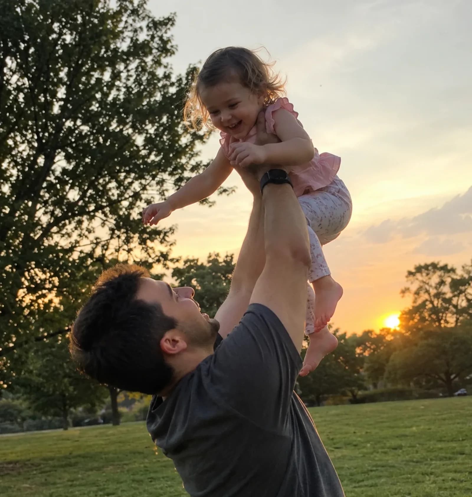 A father lifting his laughing young daughter above his head in a park at sunset